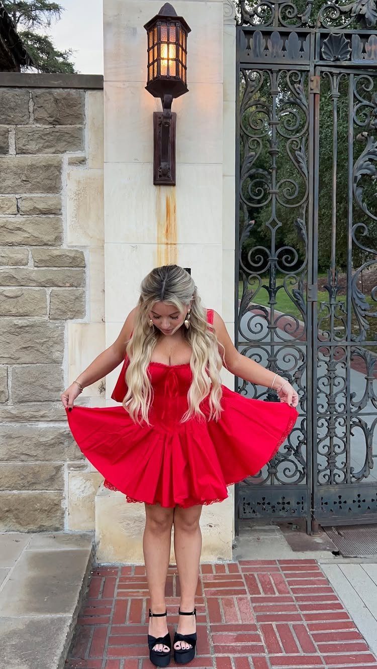 A woman in a bright red flared party dress and black platform sandals standing outdoors.
