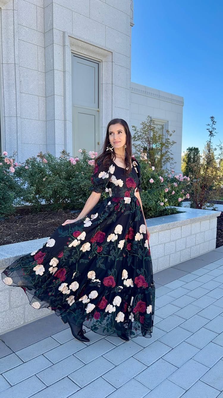 Woman posing in a long black floral maxi dress with red and white flowers outdoors.