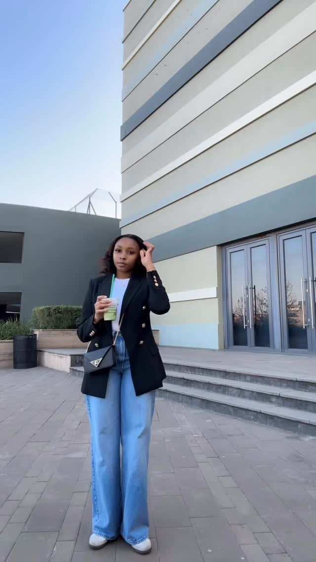 A woman posing in a black blazer, white top, and wide-leg blue jeans.
