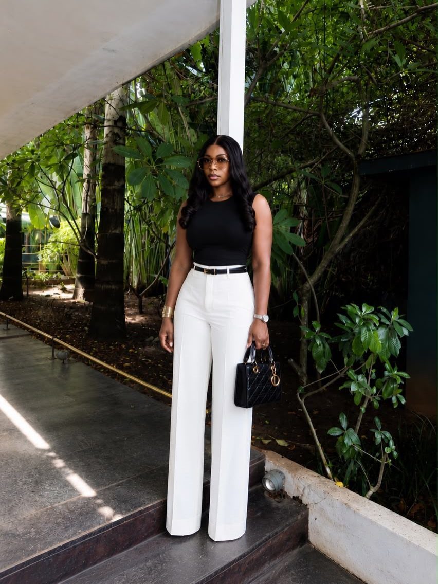 A woman posing in a black sleeveless top tucked into white wide-leg pants.