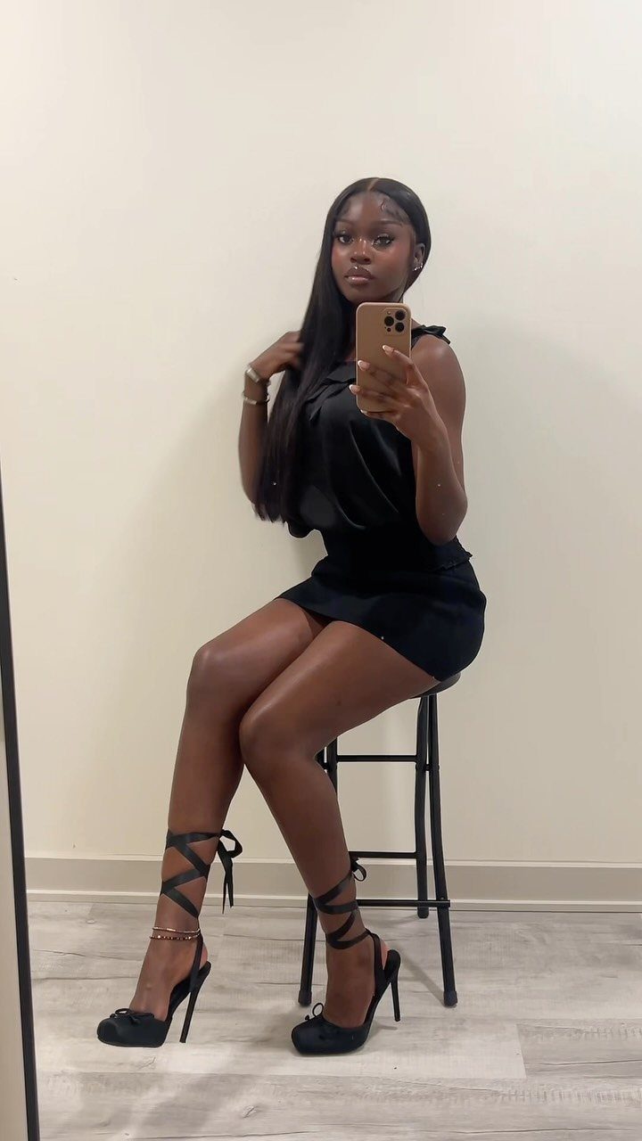 A woman in a black dress sitting on a stool wearing black strappy high heels.