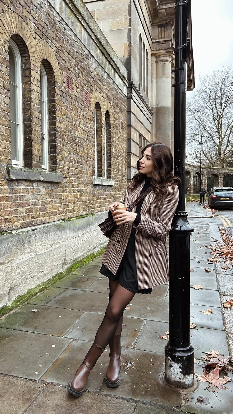 Woman in a patterned blazer and black dress wearing brown ankle boots on a city sidewalk.