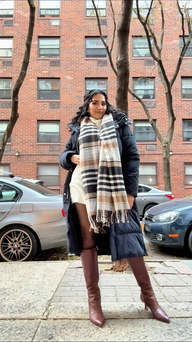 Woman posing in a dark puffer coat, plaid scarf, and brown leather knee-high boots.