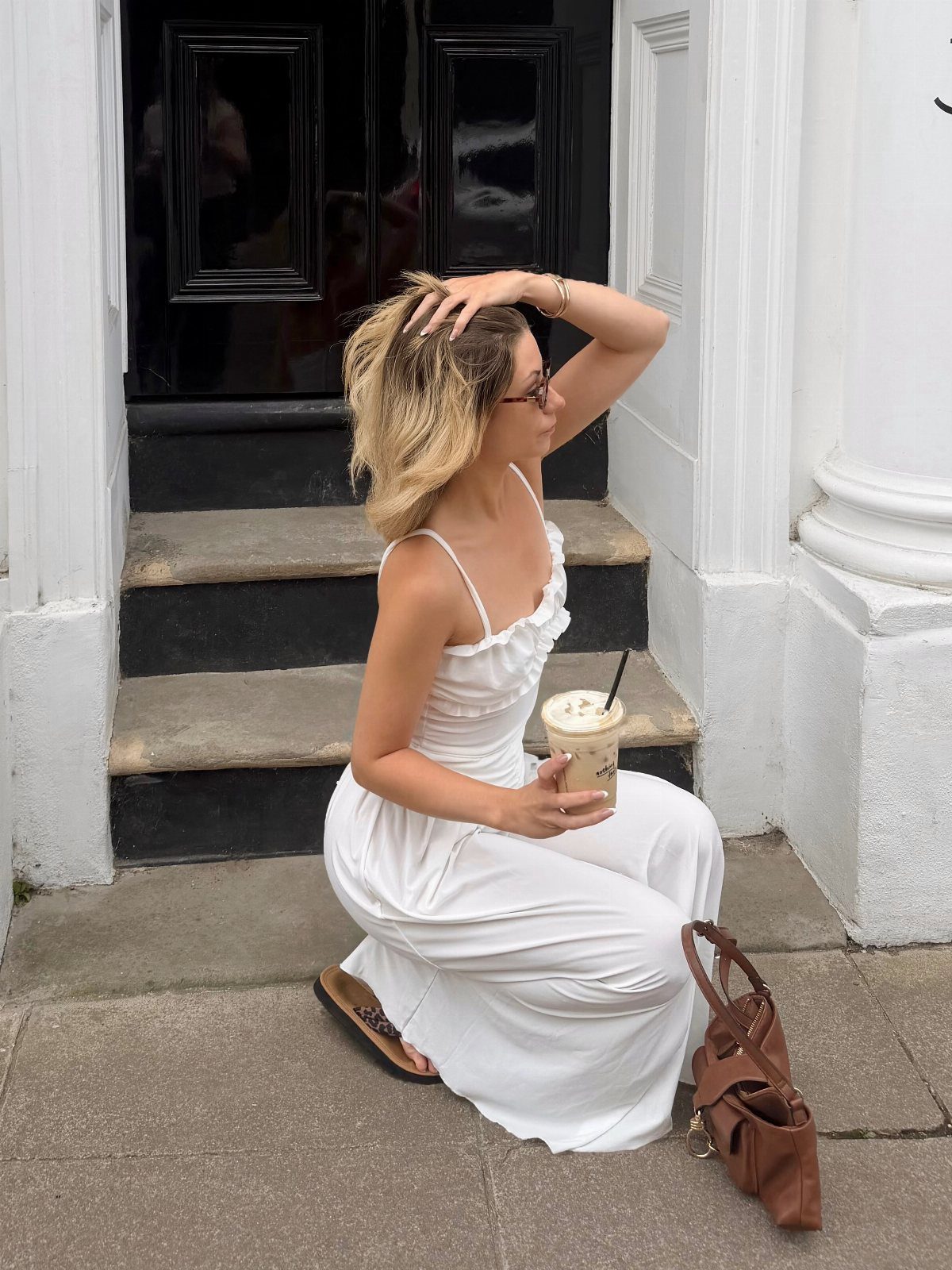 Woman in a white maxi dress and flat sandals sitting on steps with an iced coffee.