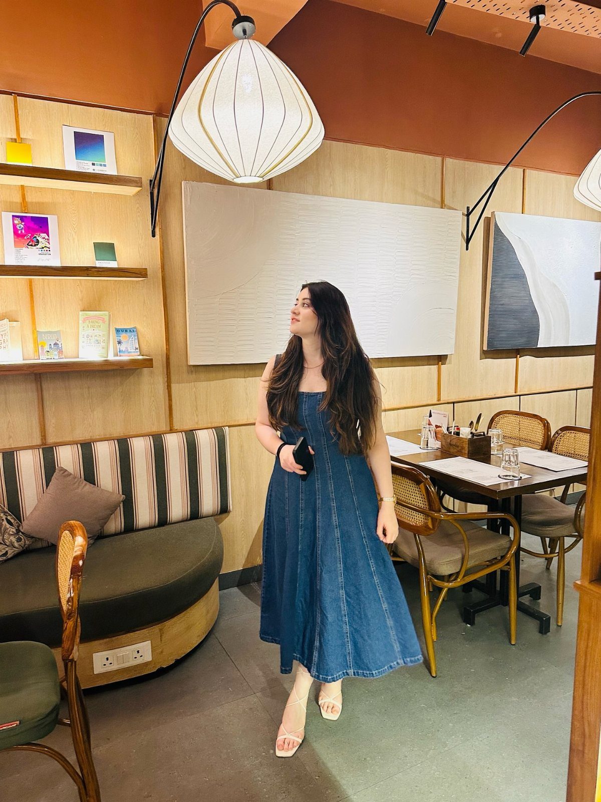 Woman in a blue denim midi sundress and white strappy sandals standing in a cafe.