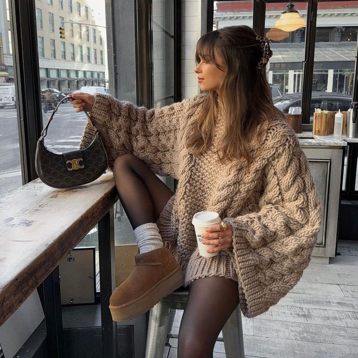 A woman in an oversized tan cable-knit sweater and platform boots sitting by a cafe window.