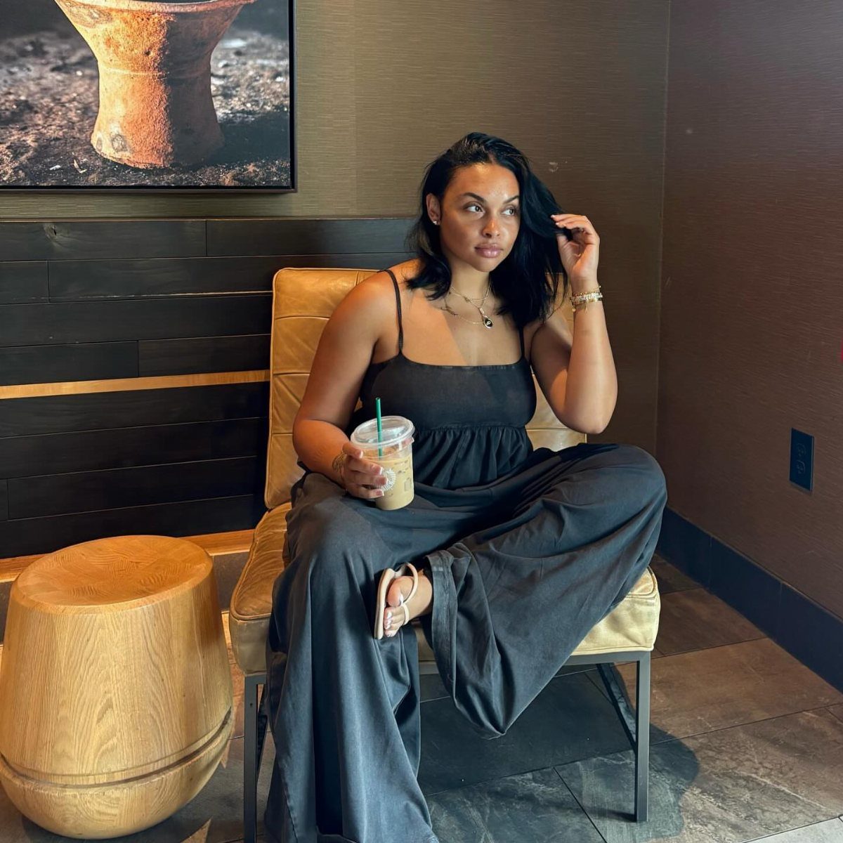 A woman in a black summer outfit sits in a cafe holding an iced coffee.
