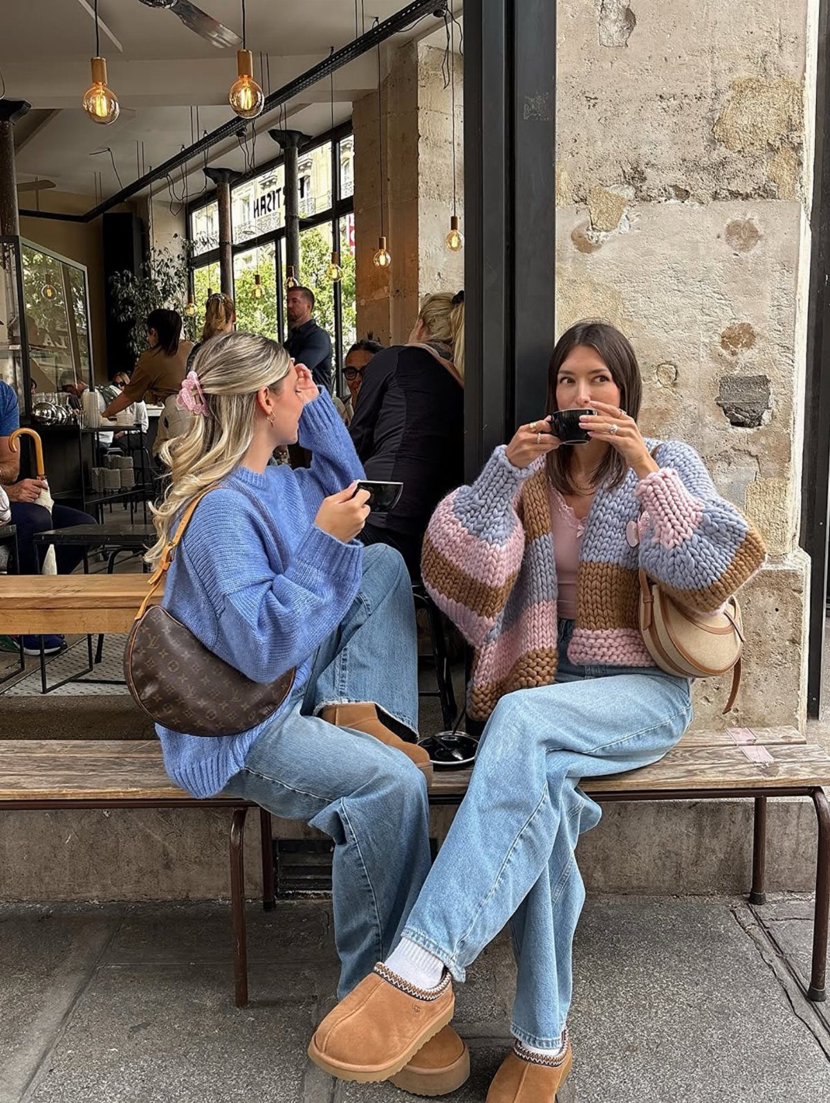 Two women sitting on a bench outside a cafe wearing chunky knit sweaters and light blue jeans.