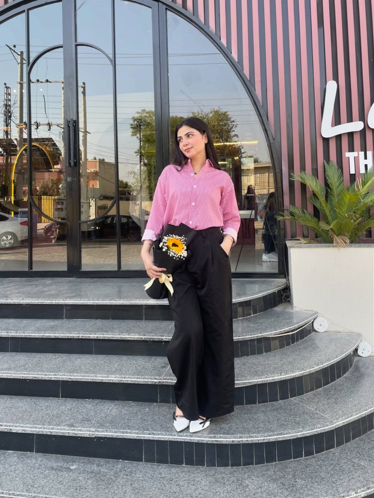Woman in a pink striped button-down shirt and black wide-leg trousers standing on outdoor steps.