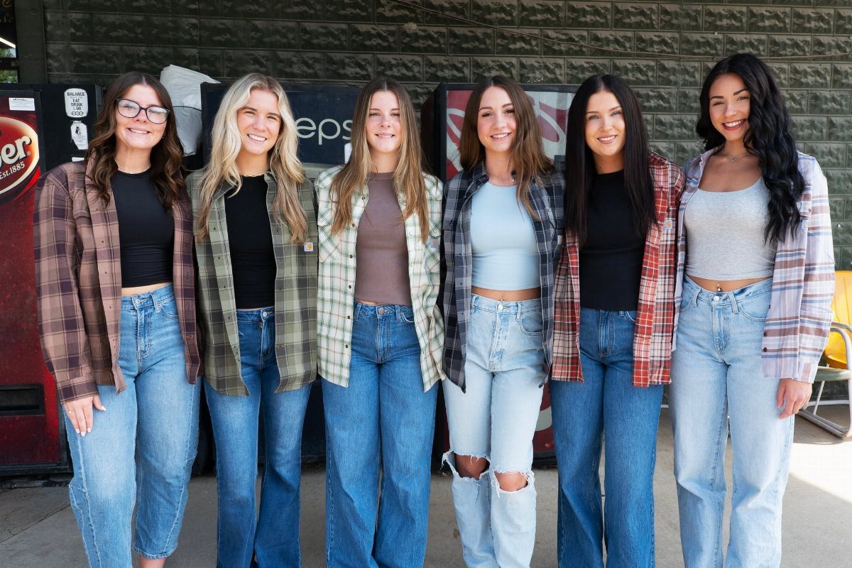Six women posing together wearing various plaid shirts and blue denim jeans outdoors.