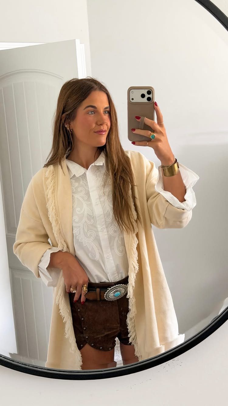 A woman posing in a mirror wearing a cream lightweight jacket, white shirt, and brown shorts.