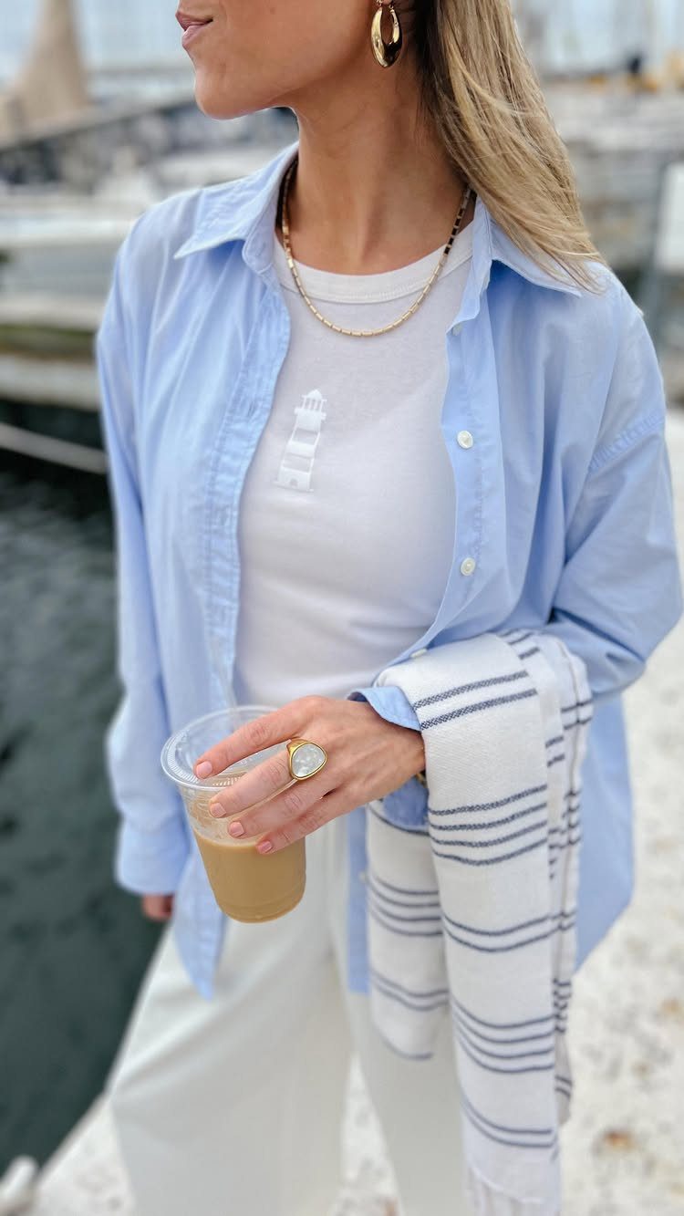 Woman wearing a light blue shirt, white t-shirt, and white trousers while holding a drink.