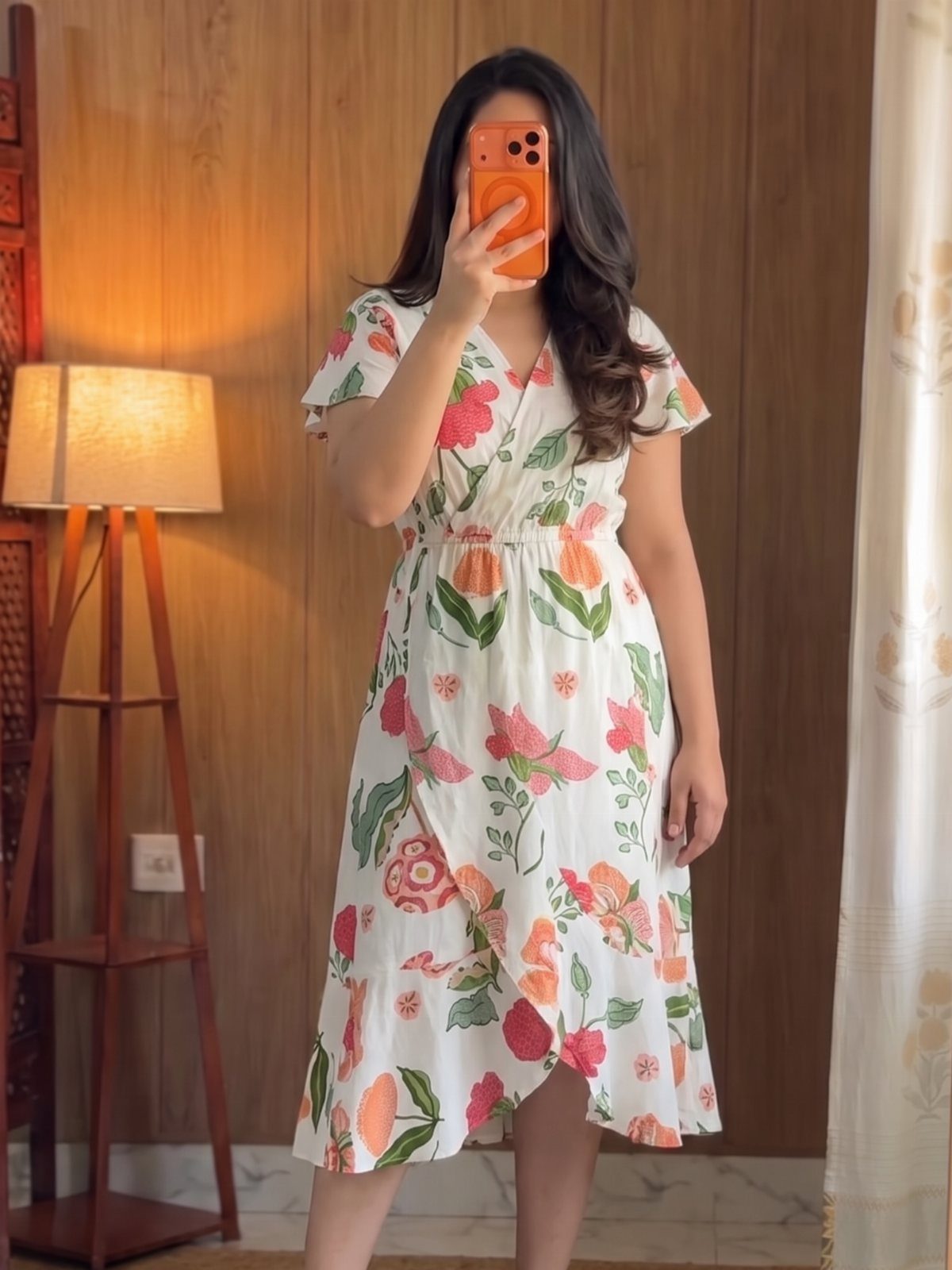 Woman in a white floral wrap sundress taking a mirror selfie in a room.