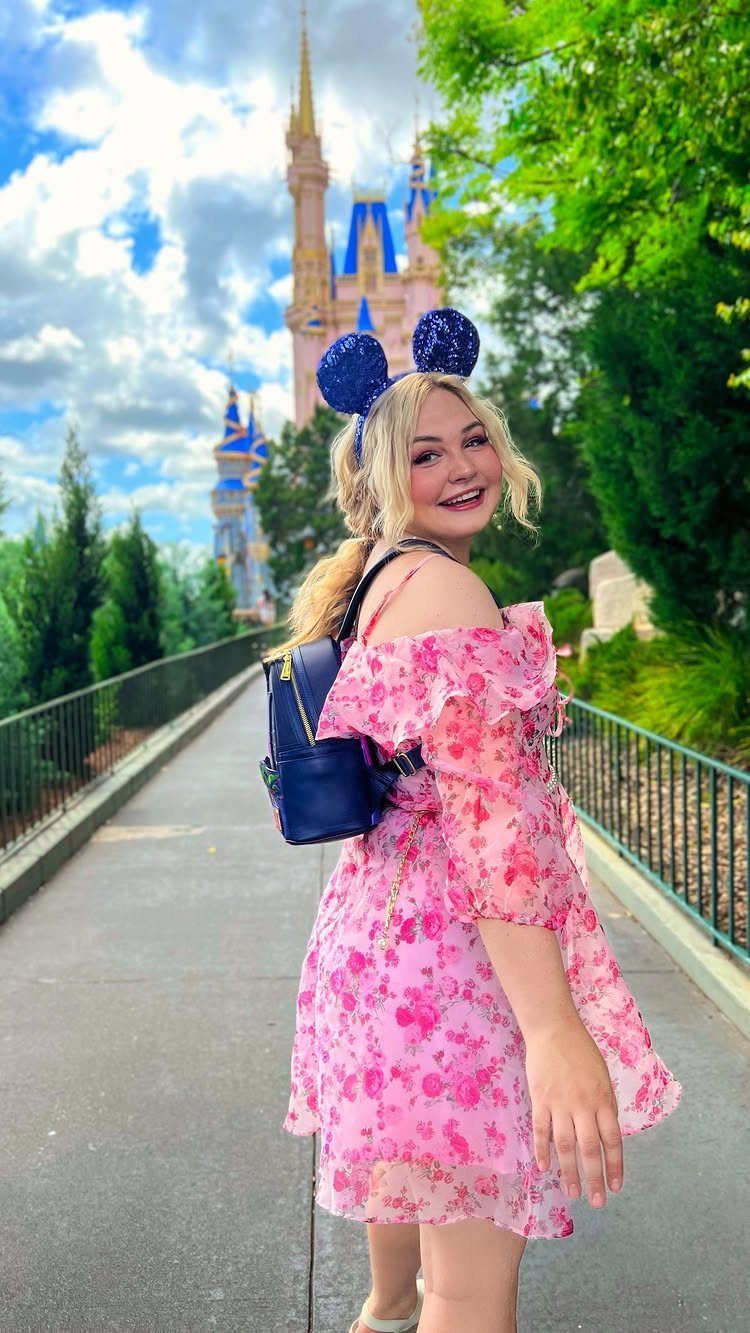 A smiling woman in a pink floral dress wearing blue glittery Minnie Mouse ears near a castle.