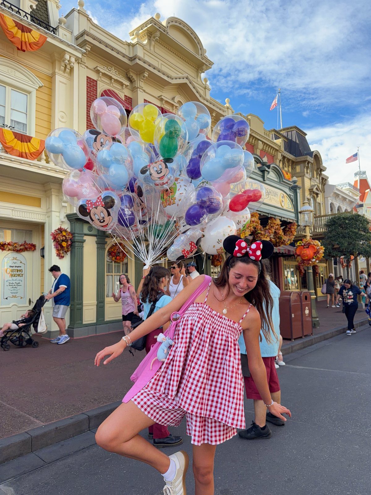 A woman wearing a gingham romper and Mickey ears holds colorful character balloons at a theme park.
