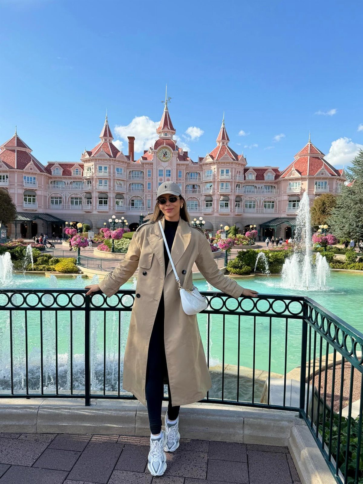 A woman in a tan trench coat and white sneakers posing by a fountain at a Disney hotel.
