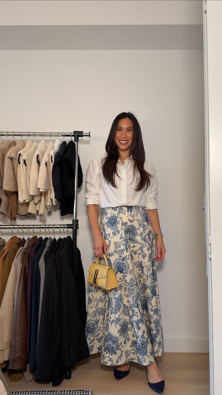 A woman smiling in a white shirt and long blue floral skirt holding a yellow purse.