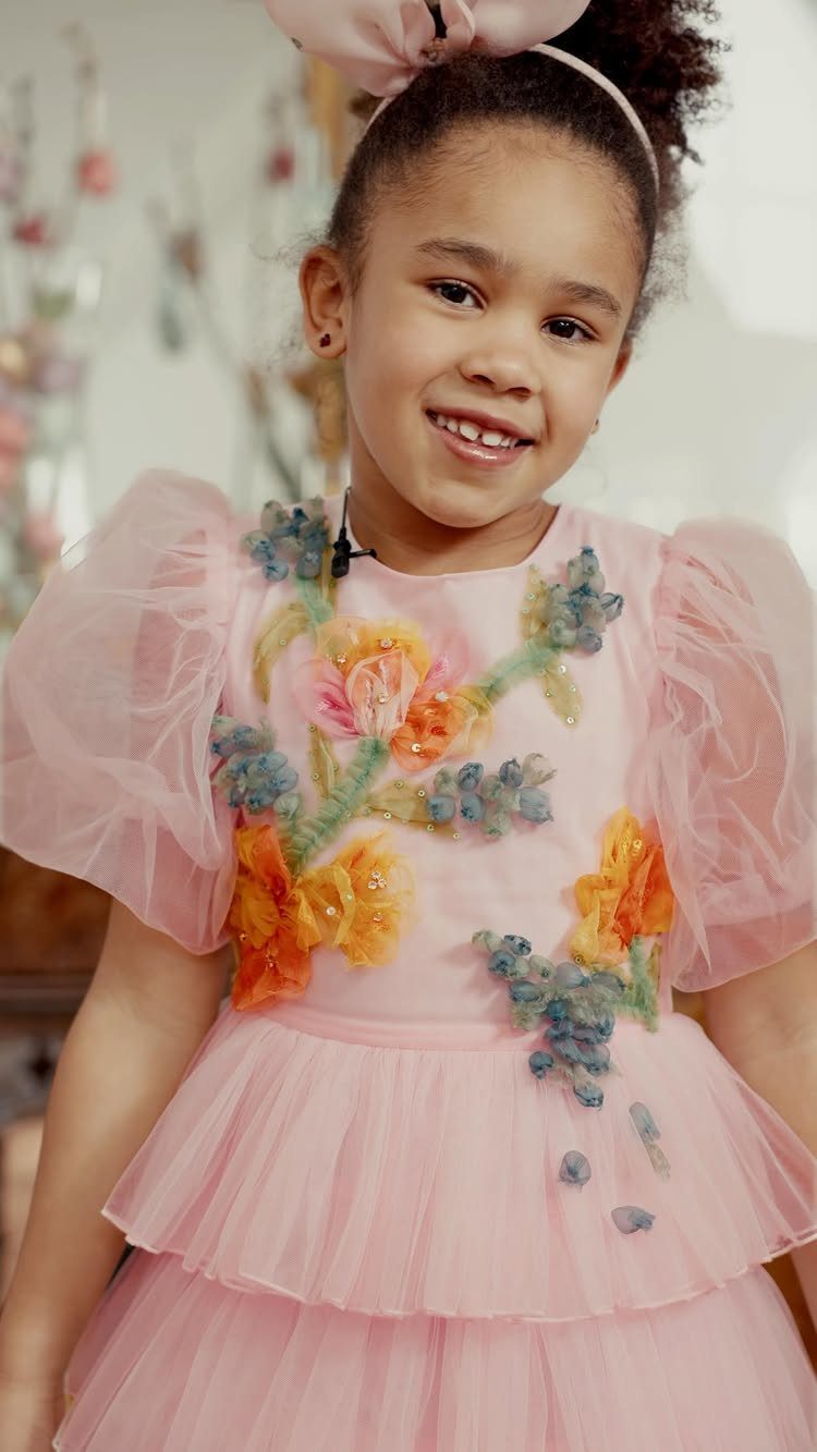 A smiling young girl wearing a pink floral embroidered tulle dress and a large hair bow.