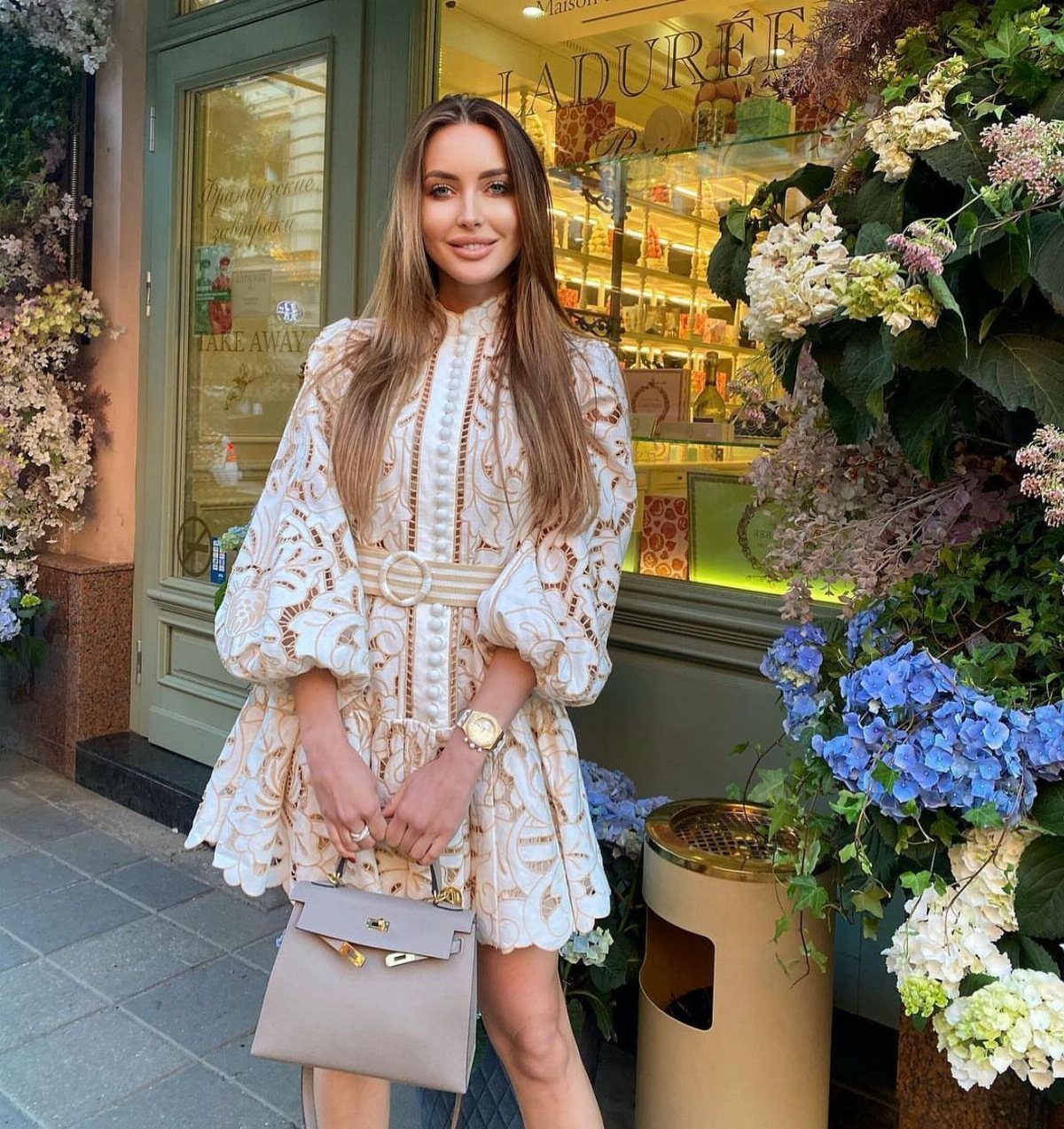 A woman wearing an embroidered white mini dress and holding a beige handbag outdoors.