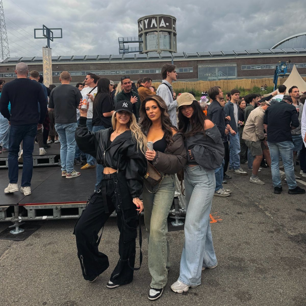 Three young women posing together at an outdoor music festival wearing casual stylish outfits.
