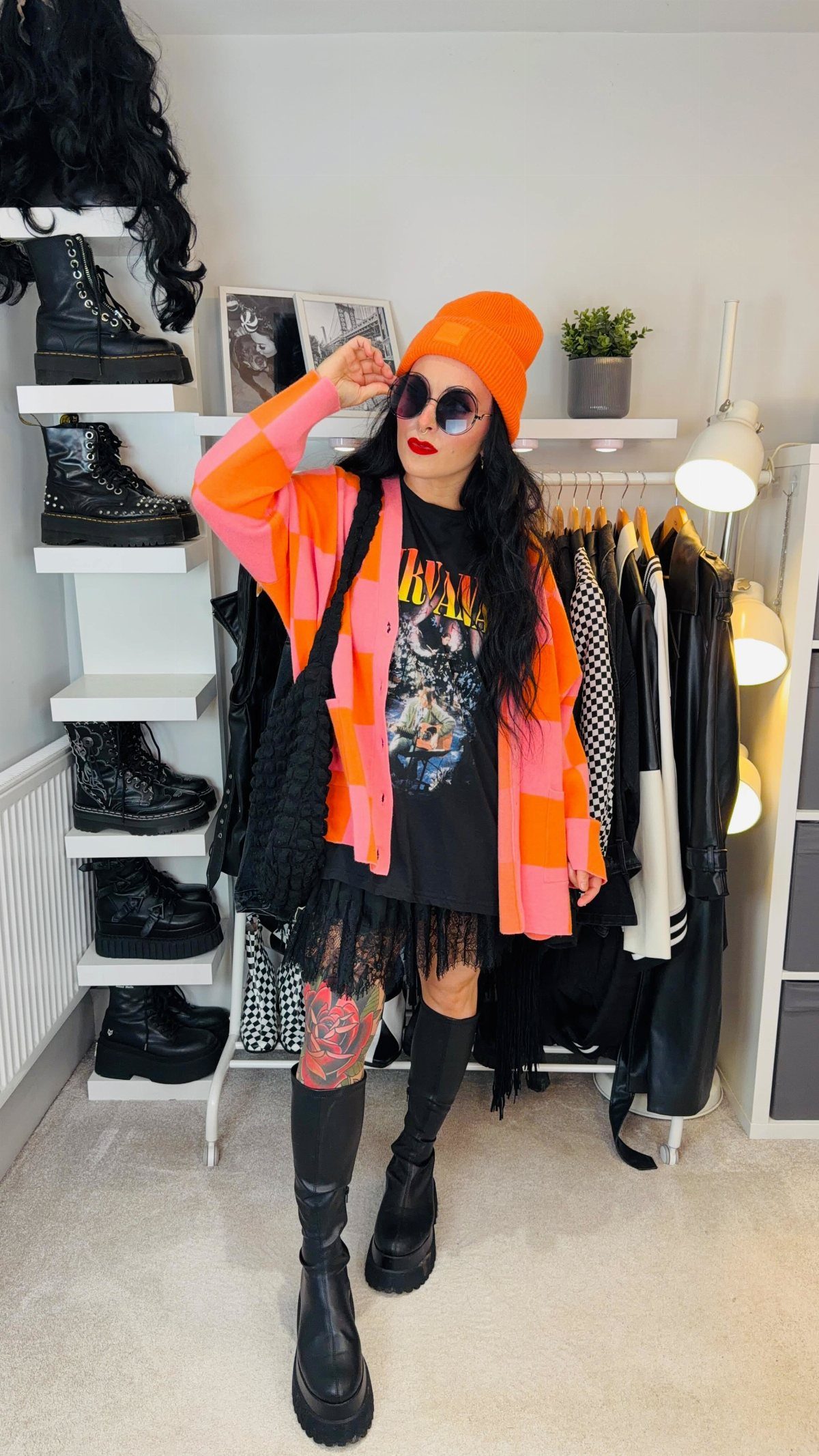 Woman in bright orange beanie and pink checkered cardigan posing in a room with clothing racks.