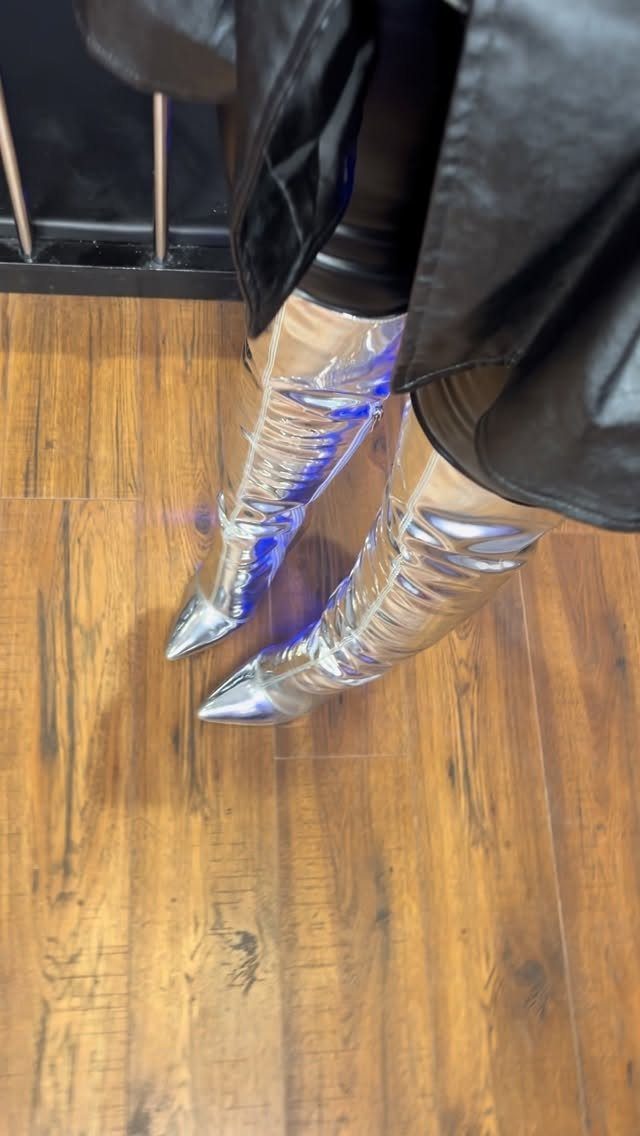 Silver metallic pointed-toe boots worn with black clothing on a wooden floor.