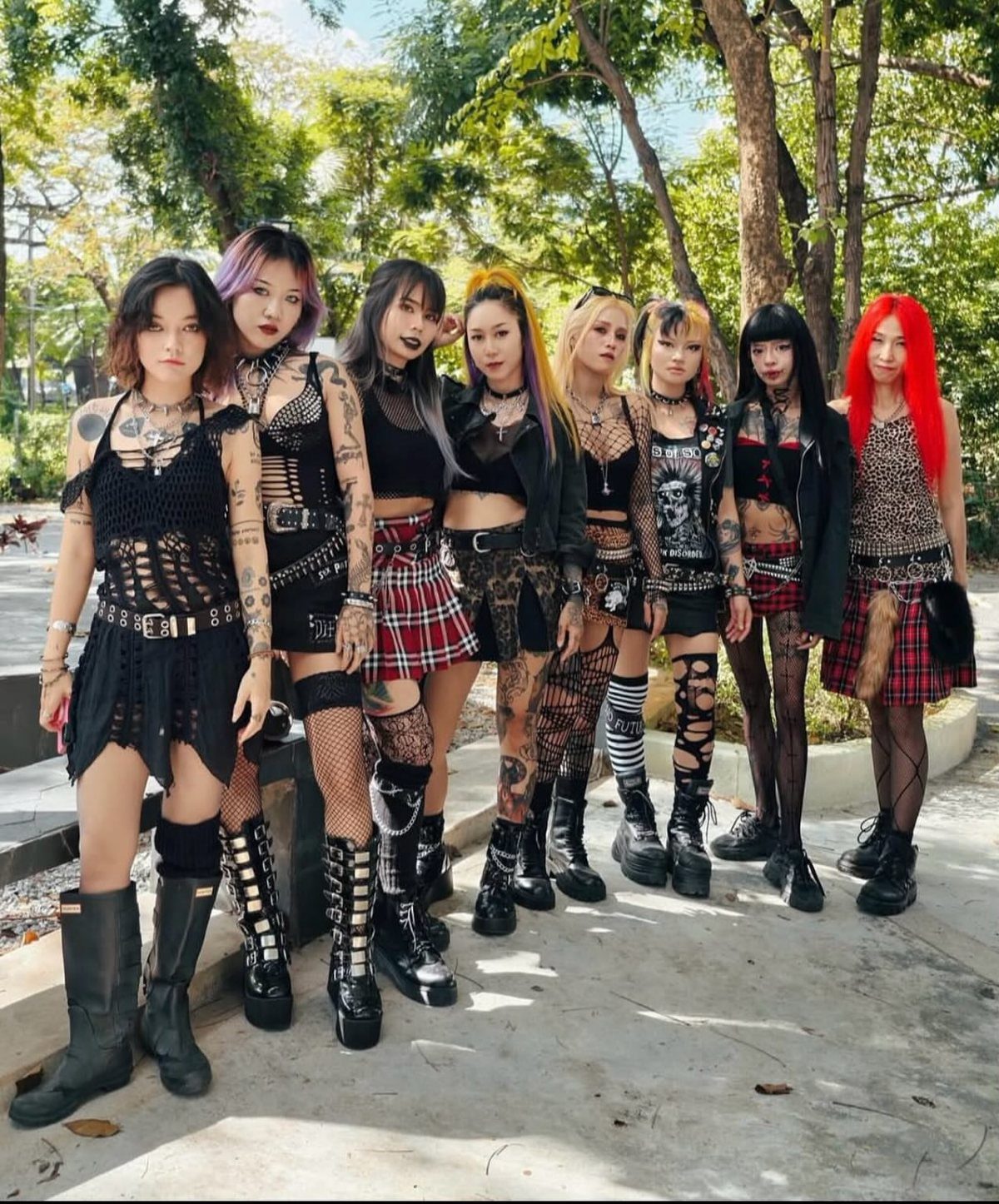Seven women in punk-style black clothing and fishnets posing together in a park.