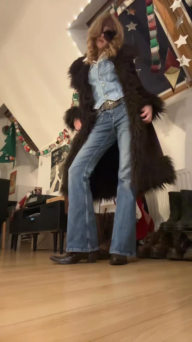 A person wearing a long dark fur coat, denim shirt, and flared bell-bottom jeans.