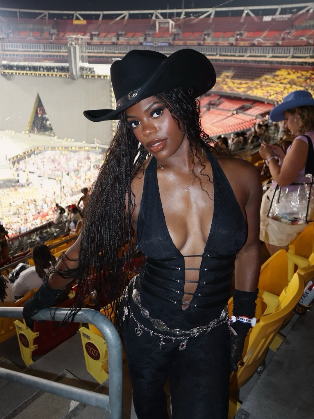 Woman in a black corset top and wide-brimmed cowboy hat posing in a stadium.