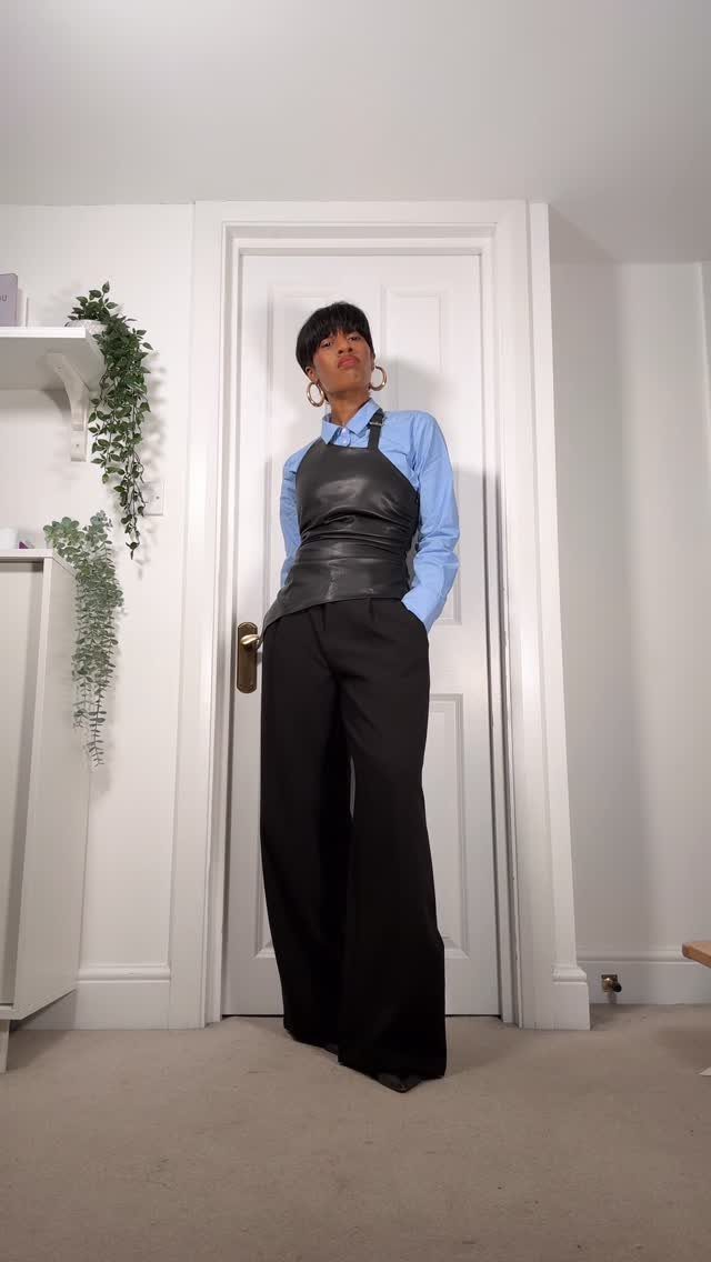 Woman wearing a blue shirt, black leather corset, and wide-leg trousers standing by a door.