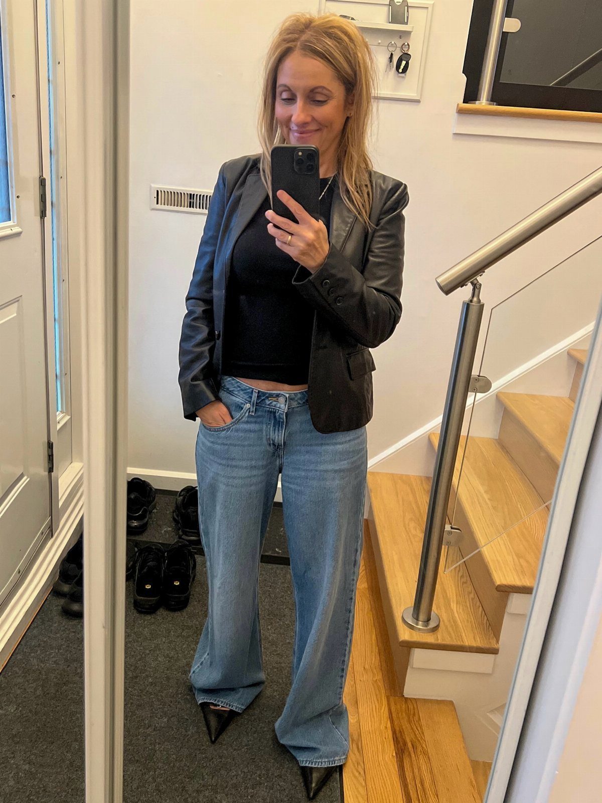 Woman in a black leather blazer and blue wide-leg jeans taking a mirror selfie.