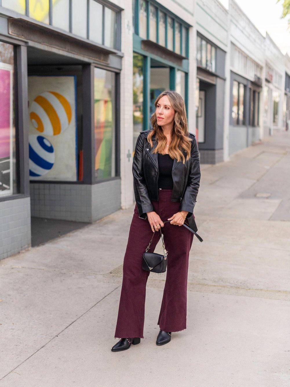 A woman posing in a black leather jacket and burgundy wide-leg trousers on a city sidewalk.