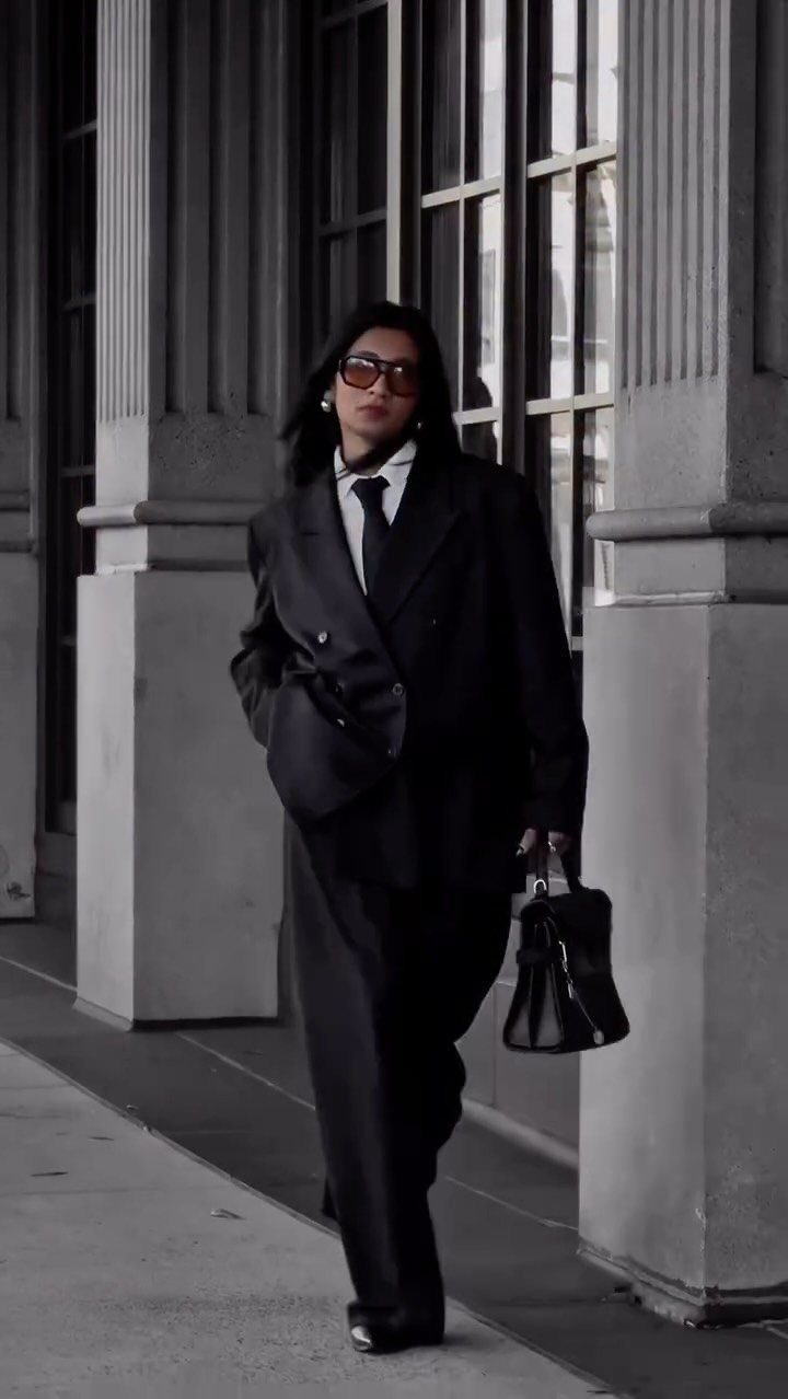 A person in an oversized black suit and sunglasses walking past large stone columns.