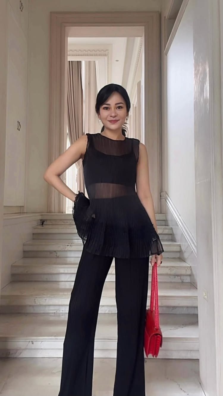 Woman in a black sheer peplum top and wide-leg trousers holding a red bag.