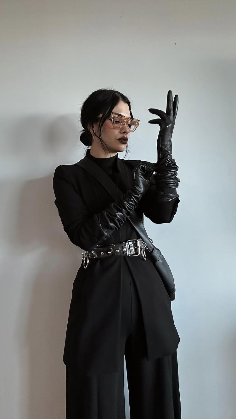 Woman in black blazer, wide-leg trousers, leather gloves, and studded belt posing against a white wall.