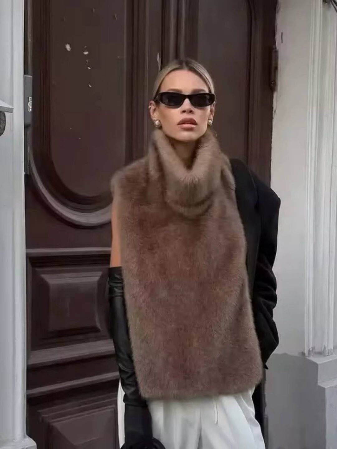 Woman in sunglasses, brown fuzzy turtleneck, and black leather gloves standing before a dark door.