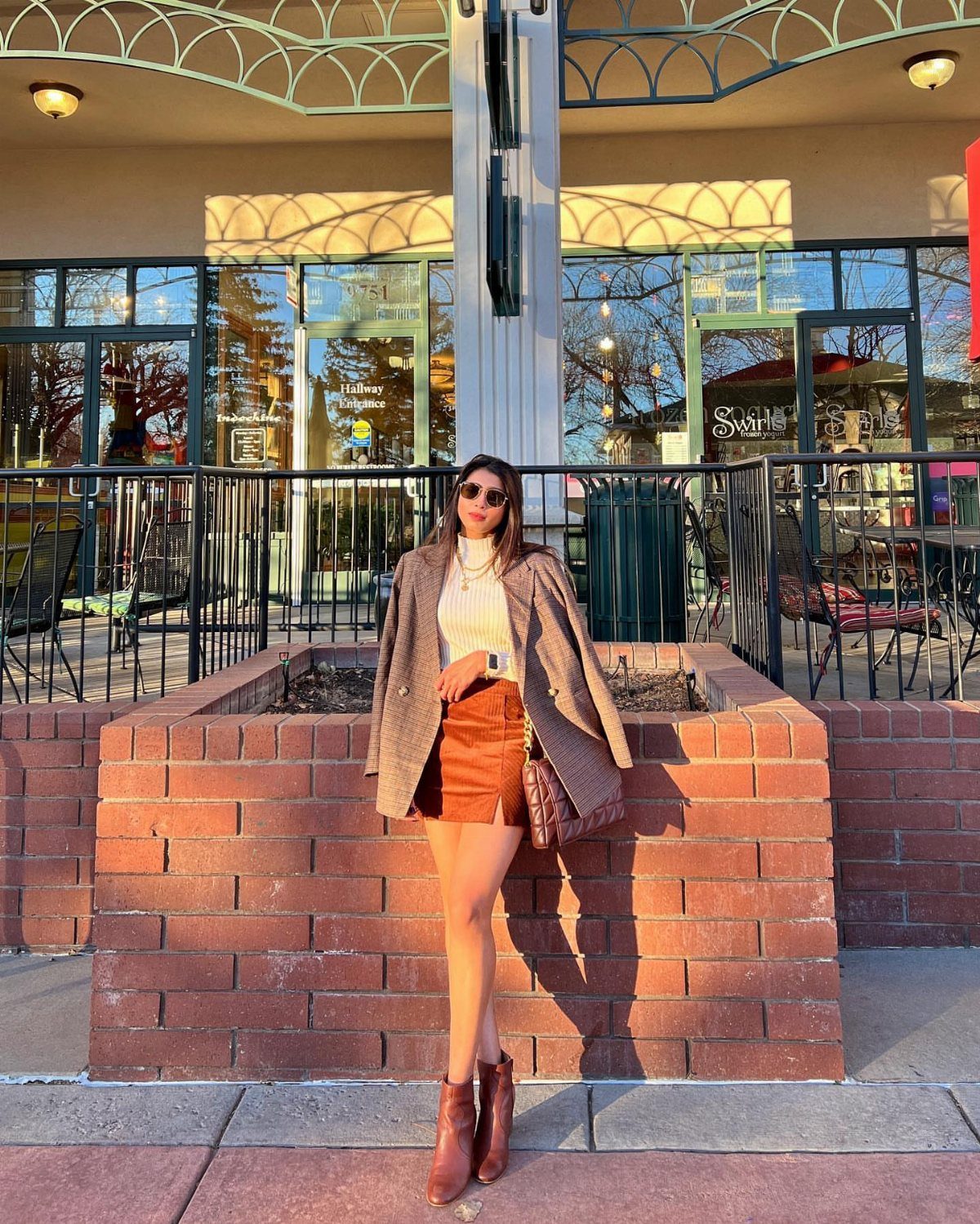 Woman in a white turtleneck, brown skirt, and blazer posing against a brick planter outdoors.