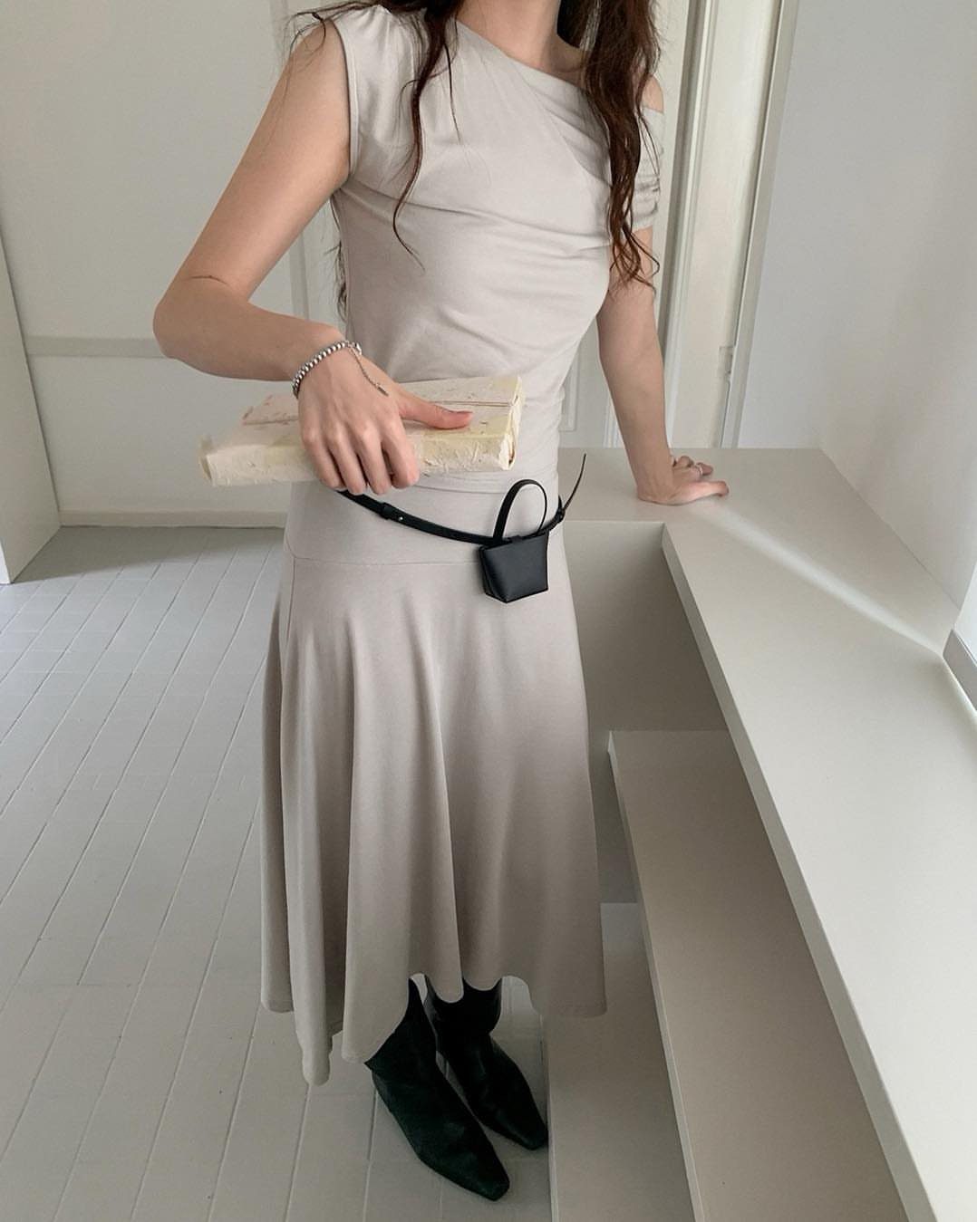 Person in a light grey draped sleeveless midi dress and black boots standing in a white room.