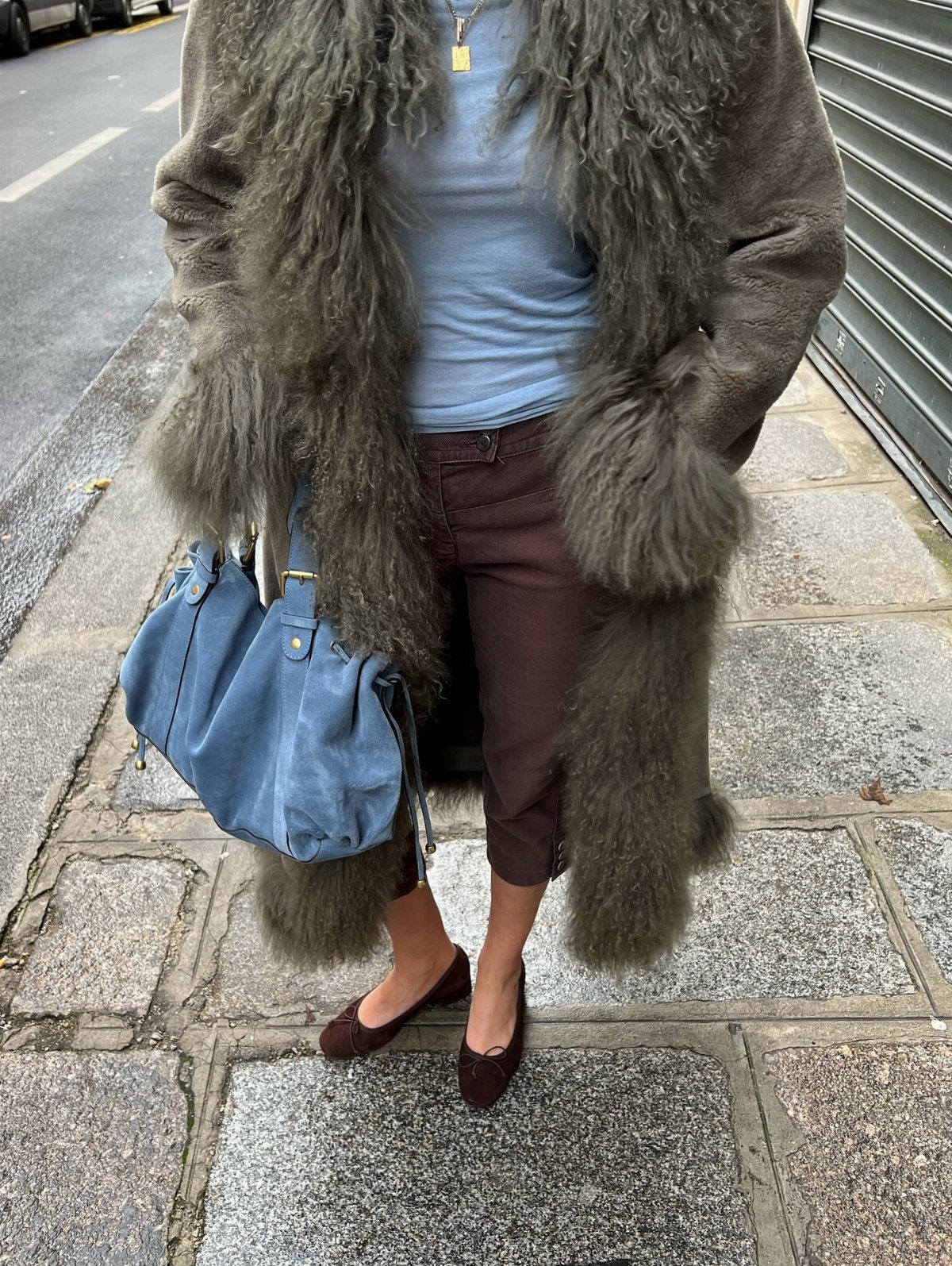 Person wearing a long olive green faux fur coat, blue top, burgundy trousers, and ballet flats.