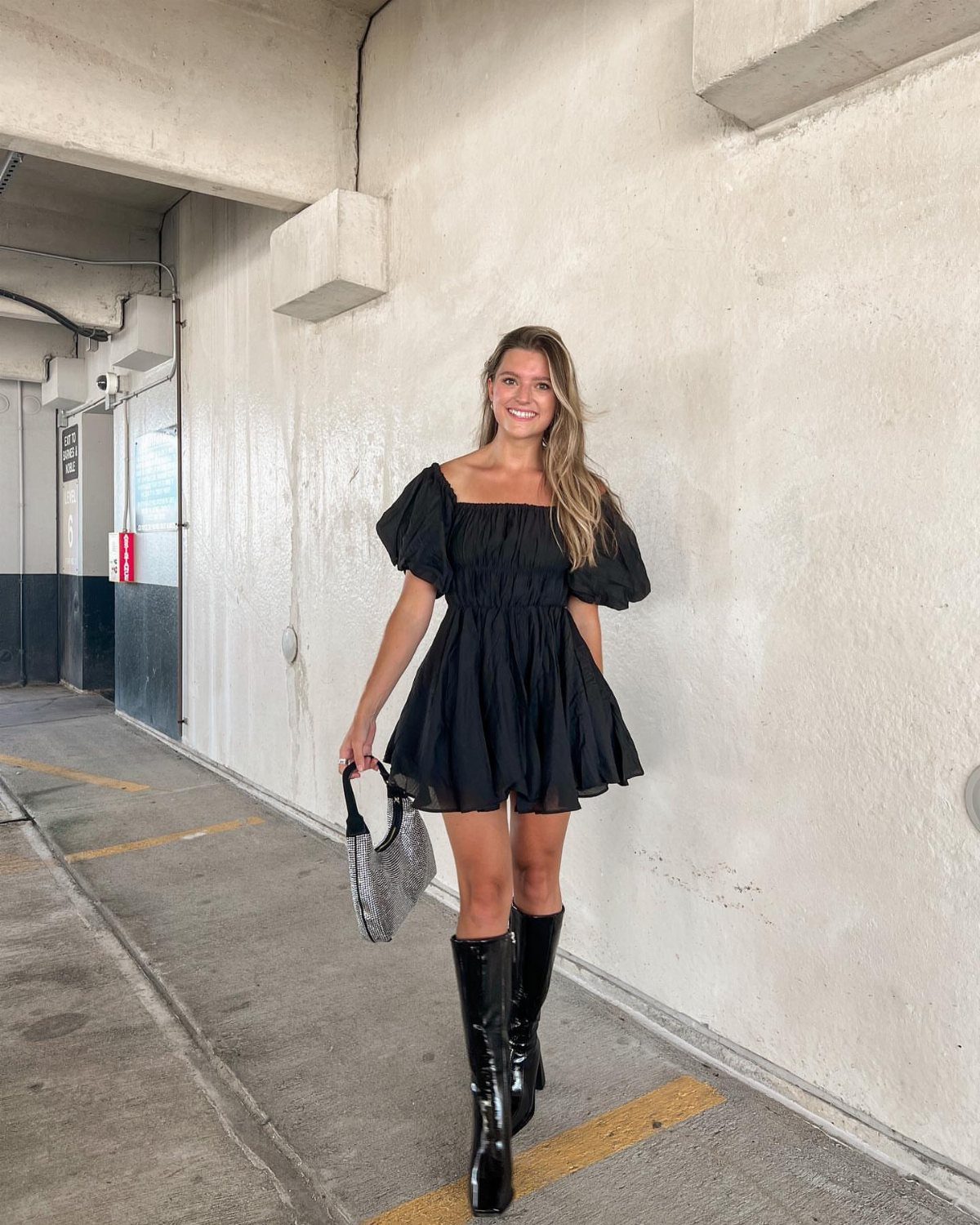 A woman wearing a black mini dress and black thigh-high patent leather boots.