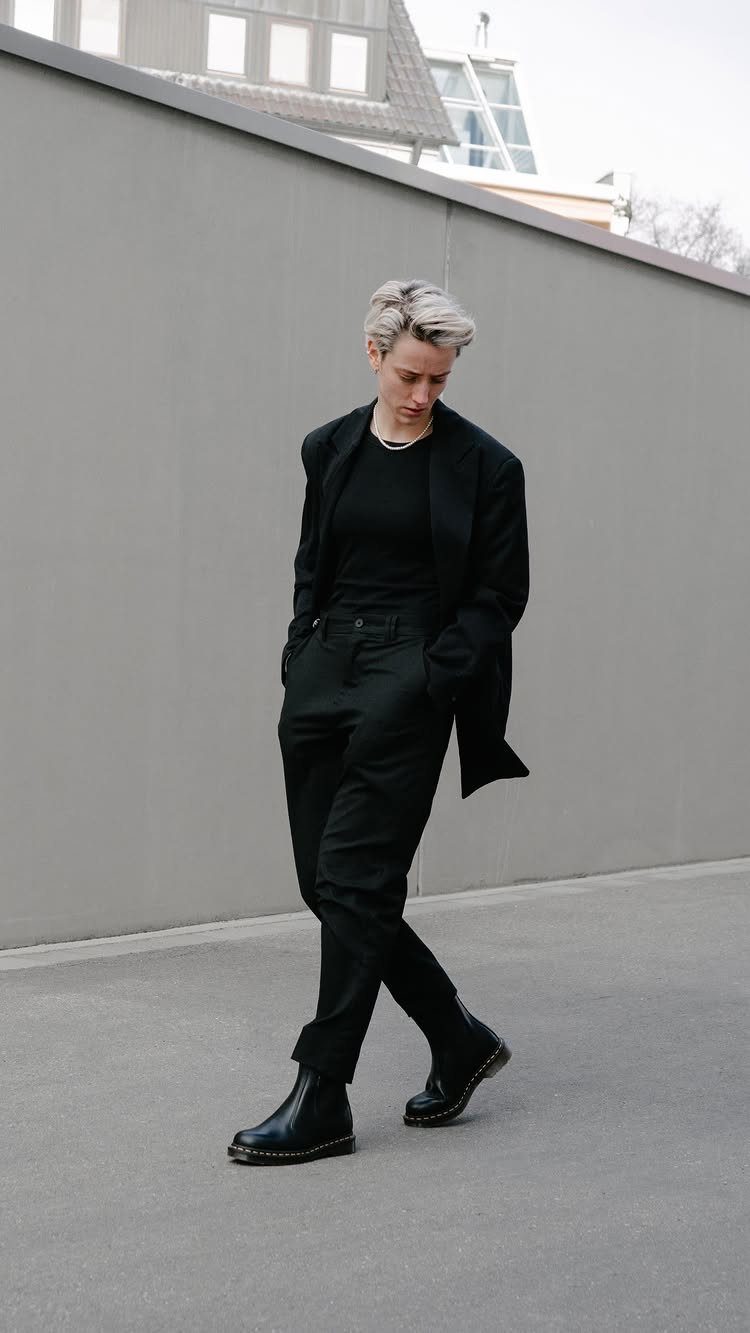 Person walking in an oversized black suit and chunky black leather boots against a grey wall.