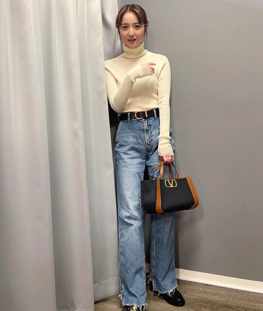 Woman wearing a cream turtleneck and wide-leg blue jeans while holding a designer handbag.