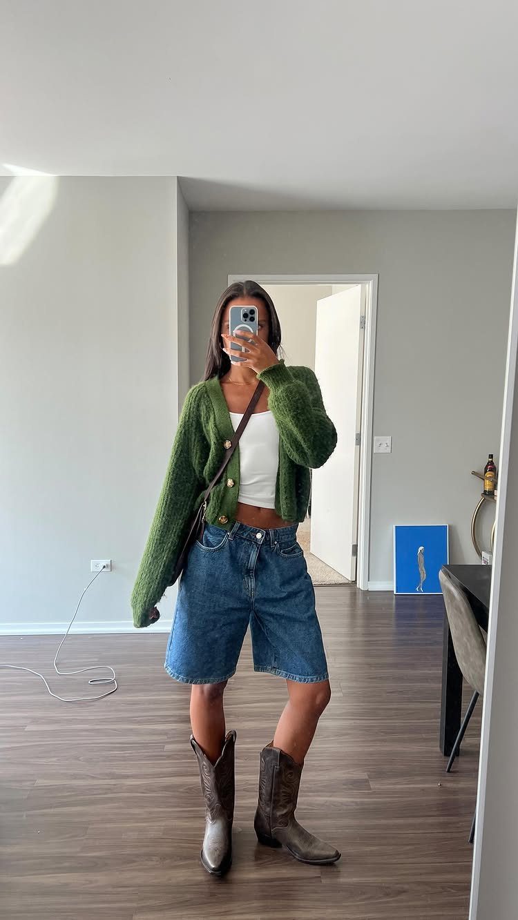 Woman in a green cardigan, white top, baggy denim shorts, and brown cowboy boots.