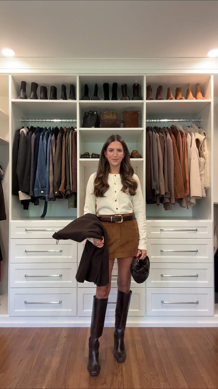 Woman in a cream cardigan, brown skirt, and tall brown boots standing in a white closet.