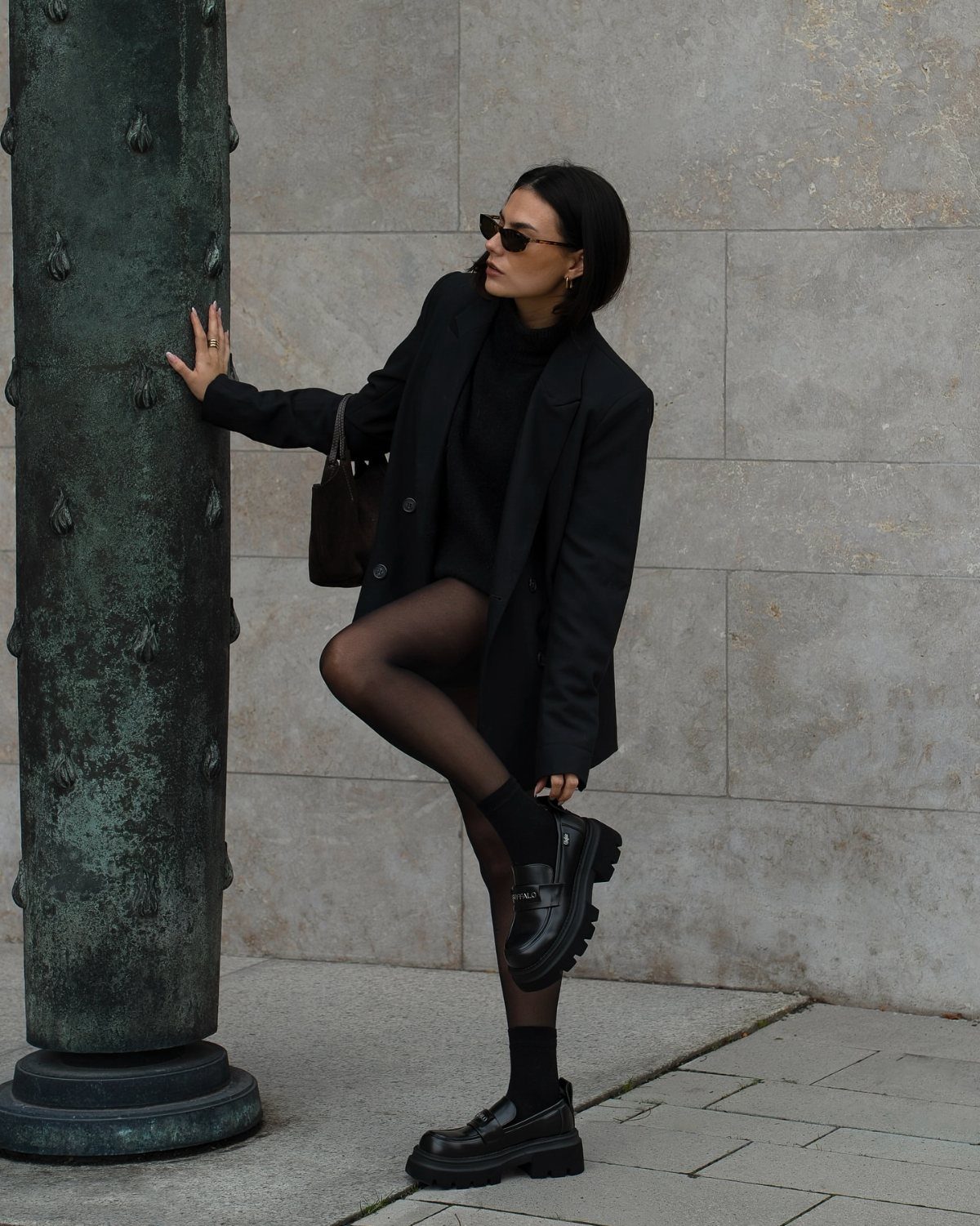 Woman in black blazer and tights wearing chunky black loafers while posing by a pillar.