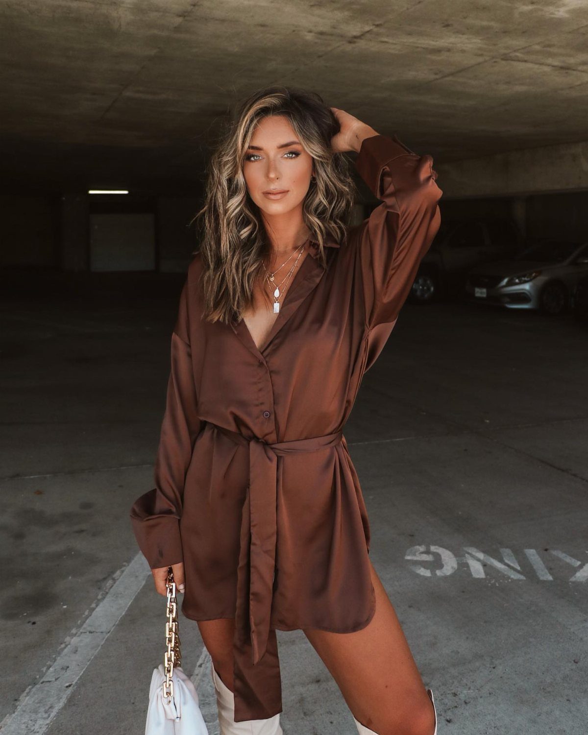 Woman in a brown satin shirt dress posing in a concrete parking garage.