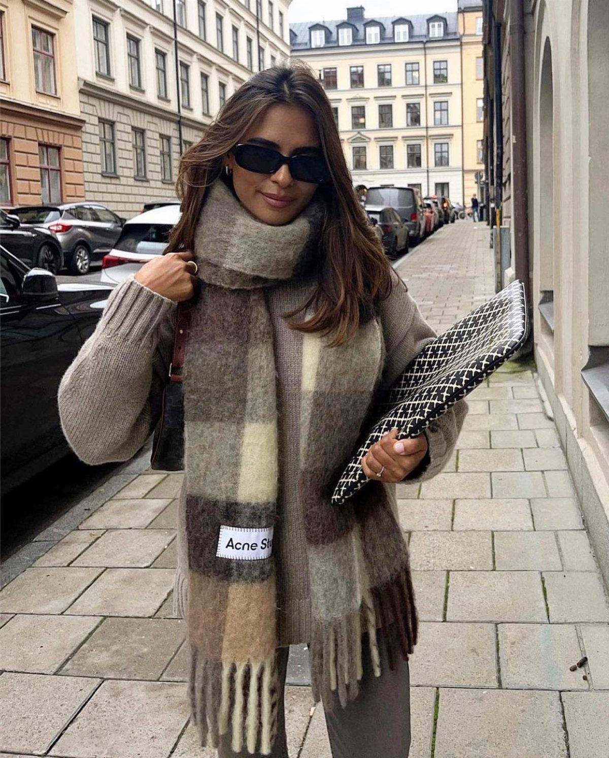 Woman wearing a large plaid scarf and oversized sweater standing on a city sidewalk.