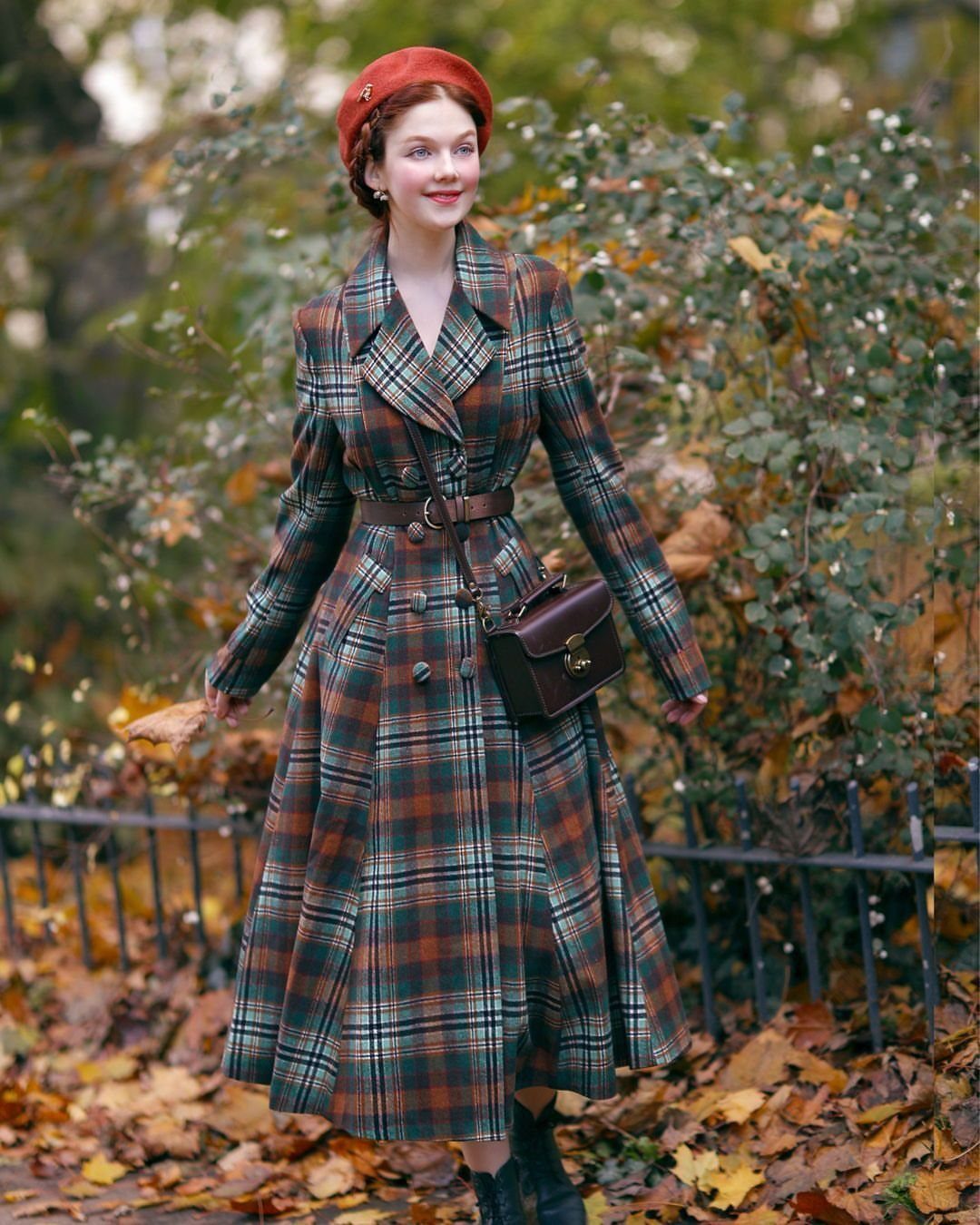 A woman in a long plaid coat and red beret standing among autumn leaves.