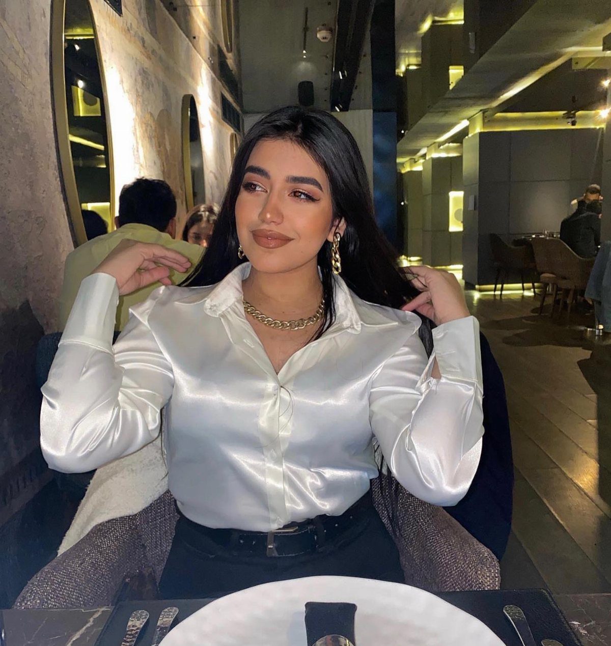 Woman wearing a white satin blouse and gold jewelry sitting at a restaurant table.
