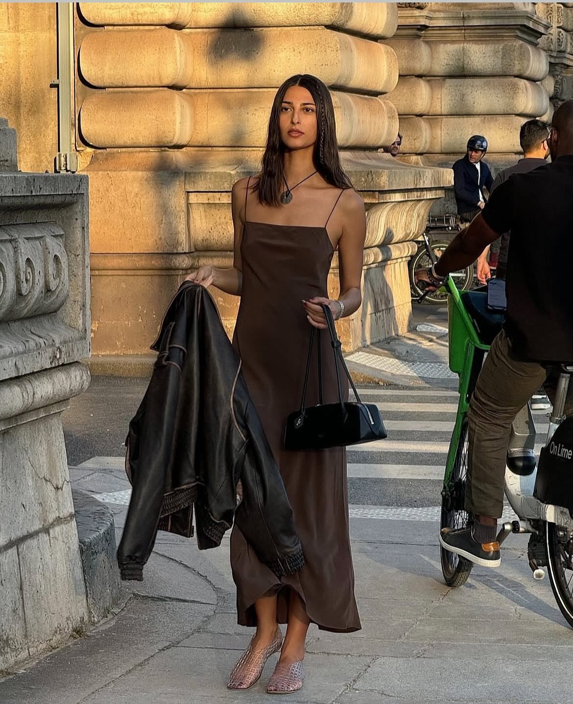 A woman wearing a brown satin slip dress walking on a city street holding a jacket.